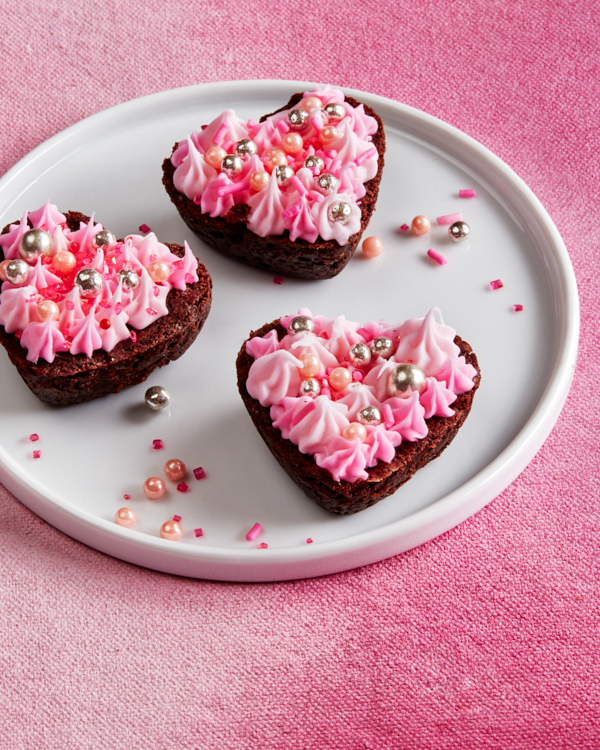 Three double chocolate brownie bites with pink icing and sprinkle decorations on a plate on a pink tablecloth.