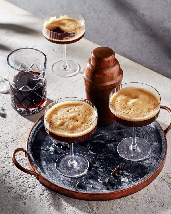 Two glasses of frothy espresso martini on a tray with a cocktail shaker with a third glass and a glass pitcher of espresso martini in the background.