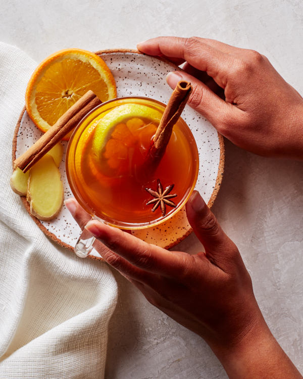 A hot toddy in a glass mug garnished with orange slices, star anise, and cinnamon sticks, on a plate with sliced ginger, cinnamon, and orange.