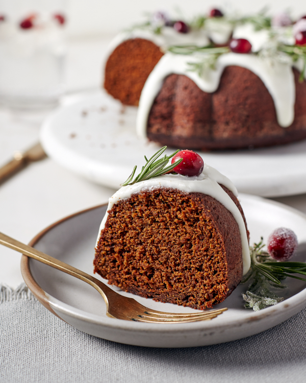 A piece of glazed gingerbread bundt cake on a plate decorated with cranberries and rosemary sprigs