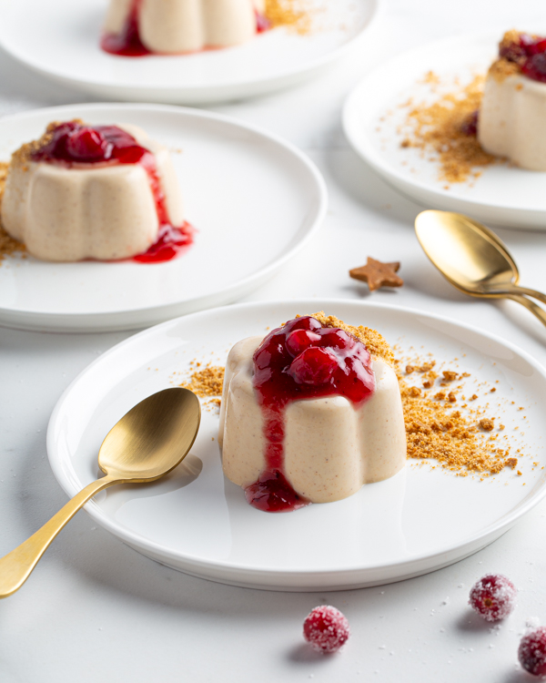 Four gingerbread buttermilk panna cottas on plates garnished with gingerbread crumbs and cranberry wine sauce