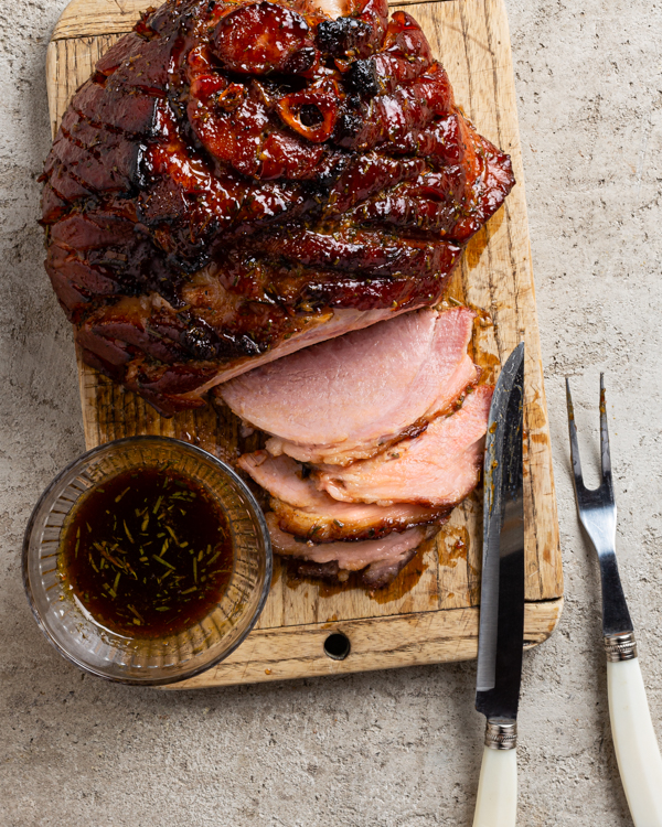 Top view of a roasted glazed ham on a cutting board with a carving knife and fork with several slices cut