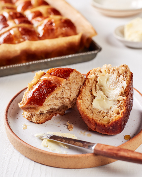 A sliced and buttered hot cross bun on a plate, with a baking sheet of hot cross buns in the background