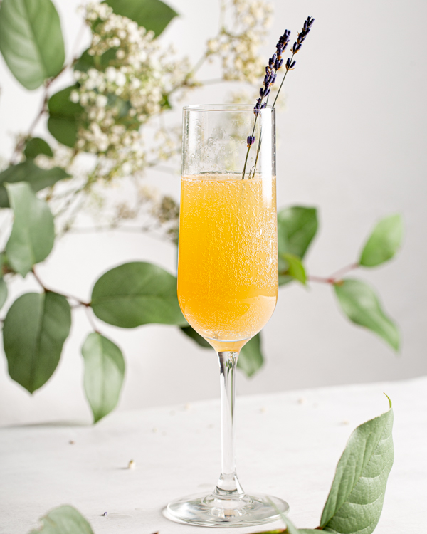 A tall glass of Lemon Lavender Mimosa garnished with fresh lavender sprigs with greenery and flowers in the background