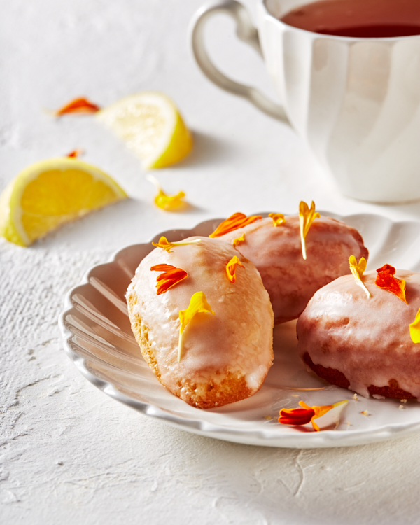 Glazed lemon madeleines decorated with edible flower petals on a saucer, shown with a cup of tea.