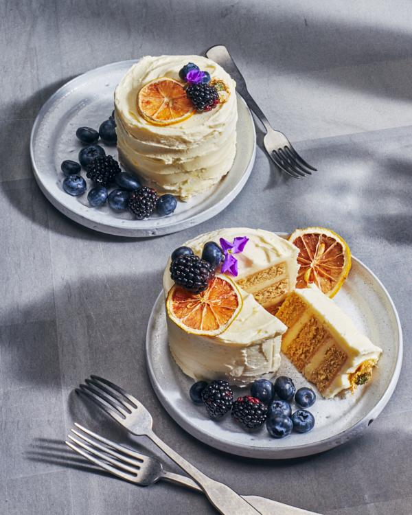 Two miniature lemon chamomile cake on plates, one with a slice cut, served with berries and topped with berries, edible flower petals, and lemon slices