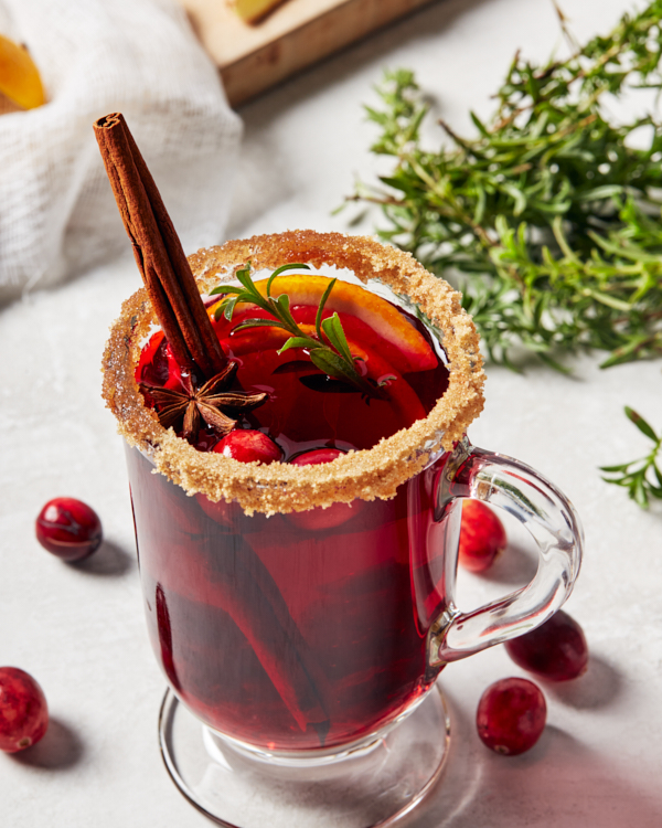 A glass mug of mulled wine garnished with a brown sugar rim, a cinnamon stick, anise, and rosemary
