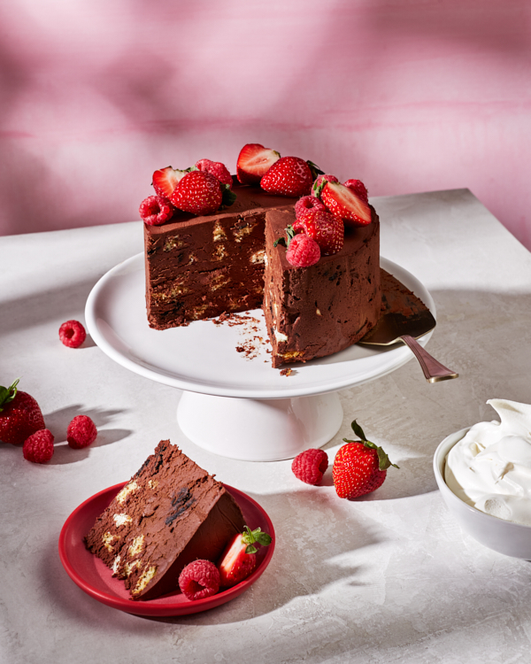 Overhead view of No-Bake Mexican Chocolate Wafer Cake topped with raspberries and strawberries, shown with two pieces cut and one on a plate, with flowers, forks, and a bowl of whipped topping.
