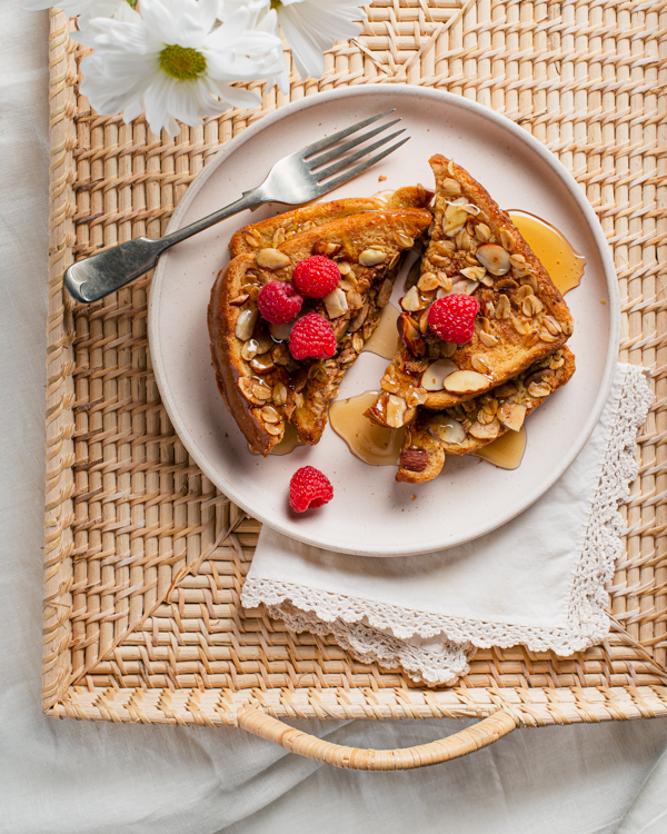 Oat and almond-crusted French toast with syrup on a rattan serving tray with a fork and flowers