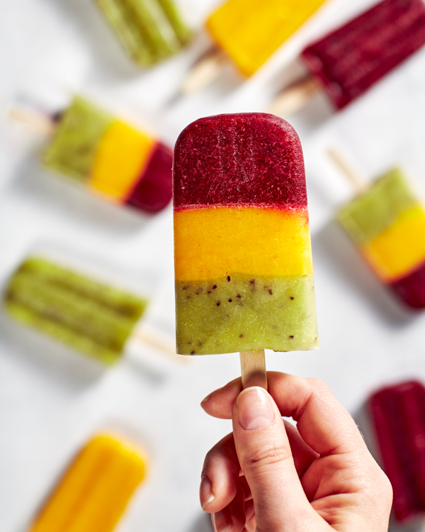A lady’s hand holding a three-layer cherry, mango, and kiwi paleta by the stick.