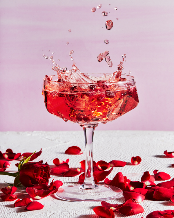 Pomegranate arils splashing into a pomegranate-rose sparkling cocktail in a stemmed glass, shown with a rose and rose petals on a table.