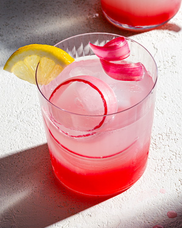 Glass of pink rhubarb lemonade garnished with a curl of rhubarb, sitting on a counter with sun shining through a window