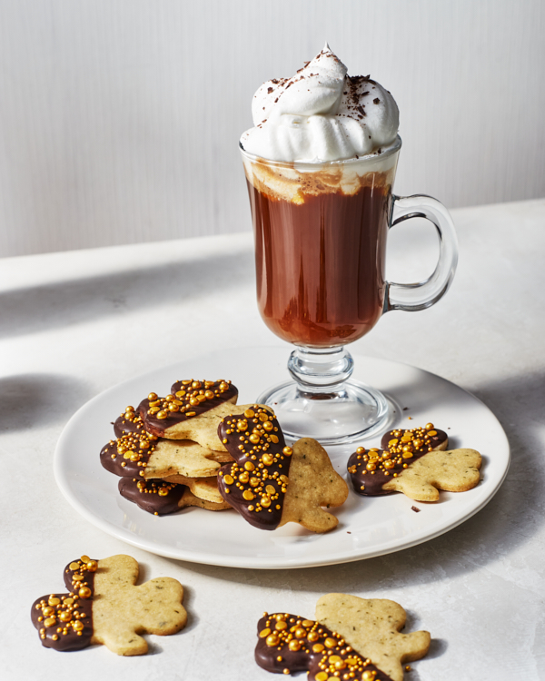 Shamrock-shaped mint cookies dipped in chocolate and decorated with gold sprinkles on a white plate served with a glass mug of hot chocolate with whipped cream.