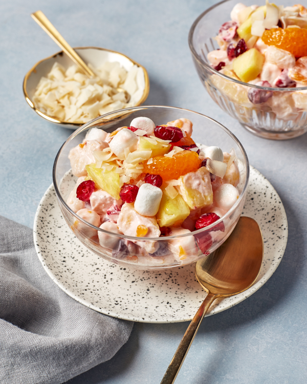 A single serving of Ambrosia salad in a glass bowl with a plate of shaved coconut shown with gold spoons