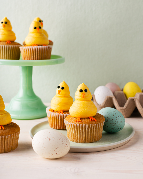 Cupcakes decorated with yellow chicks made of piped icing on a platter and a cake stand, shown with coloured eggs