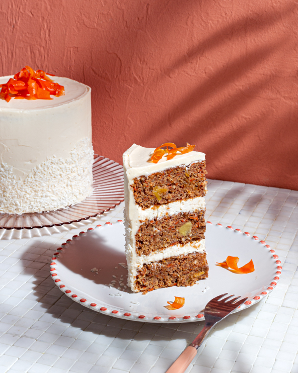A slice of three-layer vegan carrot cake topped with candied carrot curls on a plate with the full cake in the background
