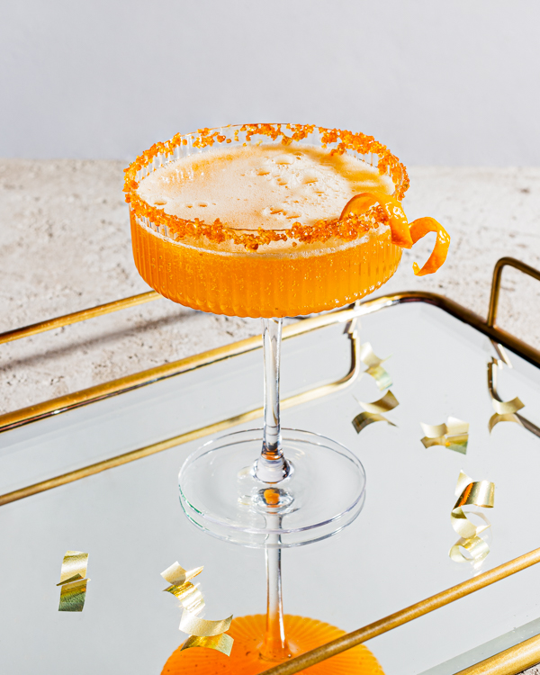  One glass of sparkling spiced ginger turmeric cocktail garnished with orange peel and rimmed with sugar on a mirrored serving tray