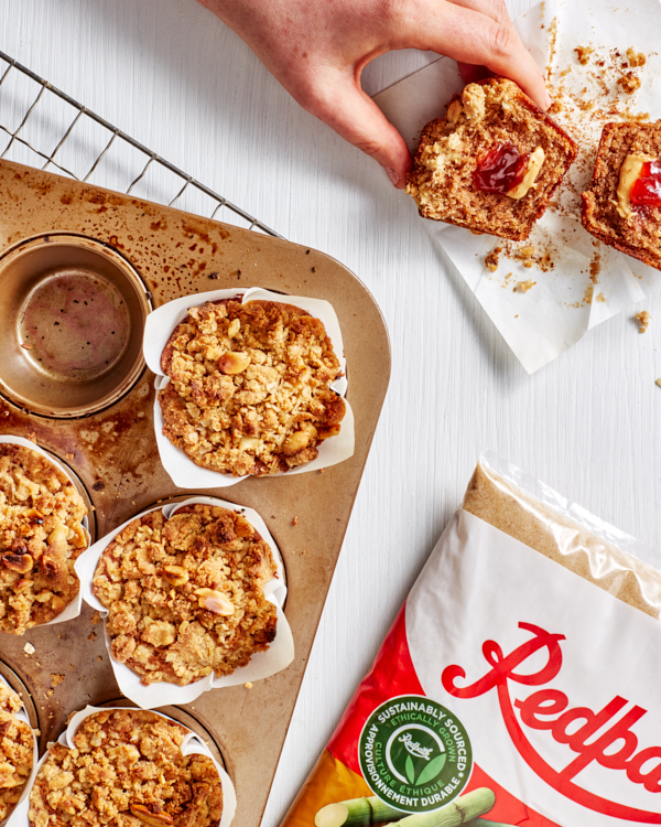 Five Vegan Peanut Butter and Jam muffins in a muffin pan, and one on a counter cut in half, shown with a bag of Redpath Golden Yellow Sugar