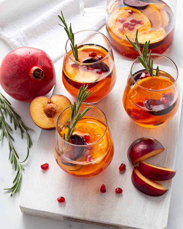 Three glasses of white sangria over ice garnished with rosemary sprigs and sliced fruit with a pomegranate
