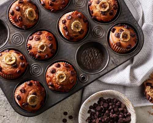 Vegan Banana Chocolate Chip Muffins in a muffin tin shown with one muffin removed and split open, and a bowl of chocolate chips