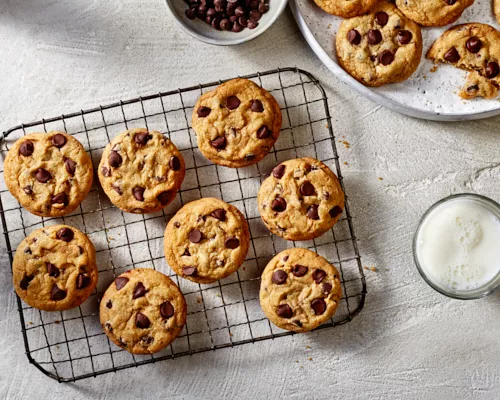 Chocolate chip cookies on a wire cooling rack with additional cookies on a platter, shown with a bowl of chocolate chips and a glass of white milk