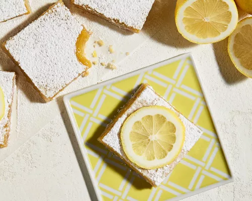 Lemon bars topped with icing sugar, two topped with a lemon slice, with one on a square plate with a yellow and white stripe pattern, shown with sliced lemons.