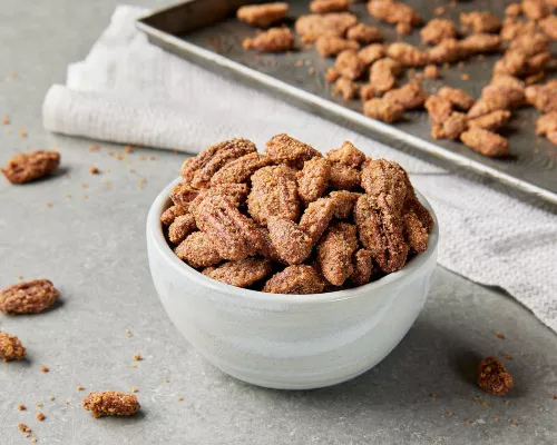 A white bowl overflowing with candied pecans and almonds, shown on a kitchen counter with a baking sheet of prepared nuts.