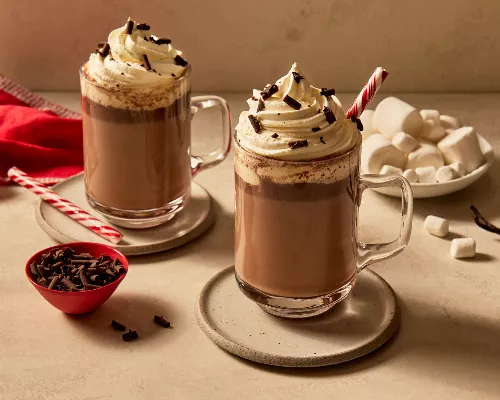 Two glass mugs of vanilla hot chocolate topped with whipped cream and garnished with shaved chocolate and candy canes, resting on saucers and shown with marshmallows, shaved chocolate, and vanilla beans. 