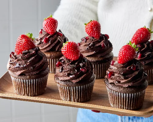 Six red wine chocolate cupcakes topped with ganache, shaved chocolate, pomegranate, and strawberries on a wooden tray held by a woman in a white sweater and jeans