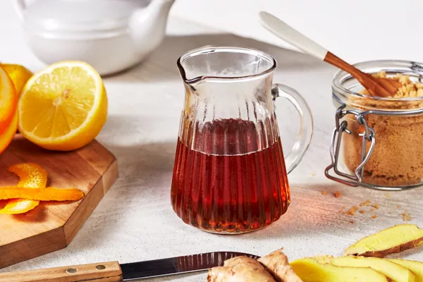 A small glass pitcher of golden brown sweet and spicy ginger syrup shown with sliced oranges and a jar of brown sugar.