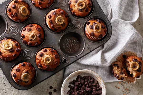 Vegan Banana Chocolate Chip Muffins in a muffin tin shown with one muffin removed and split open, and a bowl of chocolate chips.