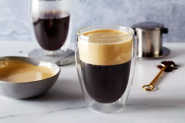 A glass of Vietnamese Egg Foam Coffee shown with a bowl of egg foam, a glass of coffee, and coffee-making equipment.