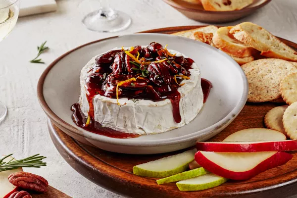 Baked brie topped with spiced candied pecans and Dark Brown sugar syrup and garnished with orange zest and rosemary, shown on a plate on a serving board with crackers, crostini, and sliced pears and apples.