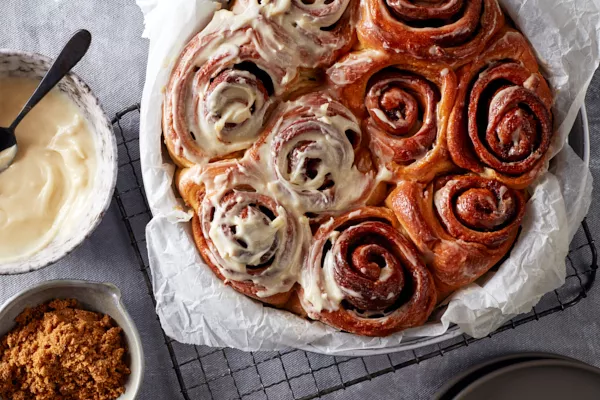 A round baking dish lined with parchment and full of cinnamon rolls, half of them iced, shown with a bowl of icing and a bowl of Dark Brown sugar