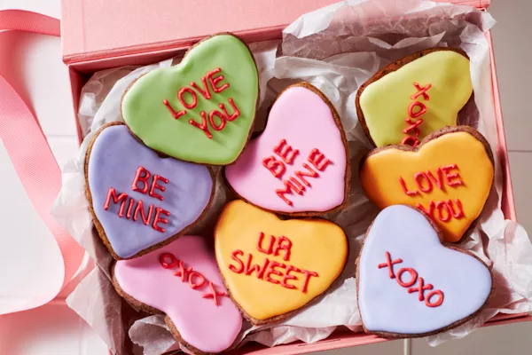 Heart-shaped sugar cookies decorated with royal icing with romantic messages written in icing, shown on white tiles with bowls of royal icing and a piping bag.