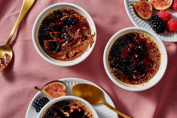 Three ramekins of salted dark chocolate crème brûlée, one partially eaten, shown with gold spoons, figs, and berries, on a rose-coloured tablecloth.