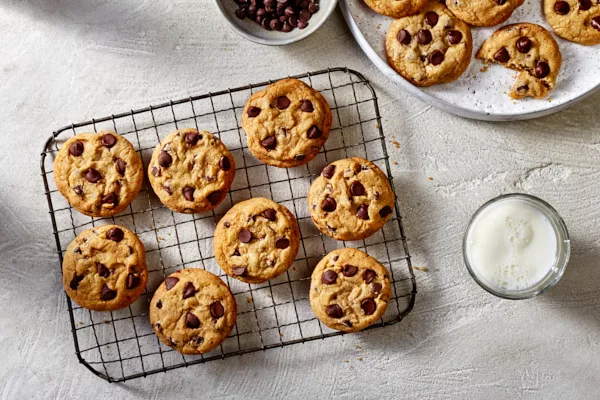 Chocolate chip cookies on a wire cooling rack with additional cookies on a platter, shown with a bowl of chocolate chips and a glass of white milk