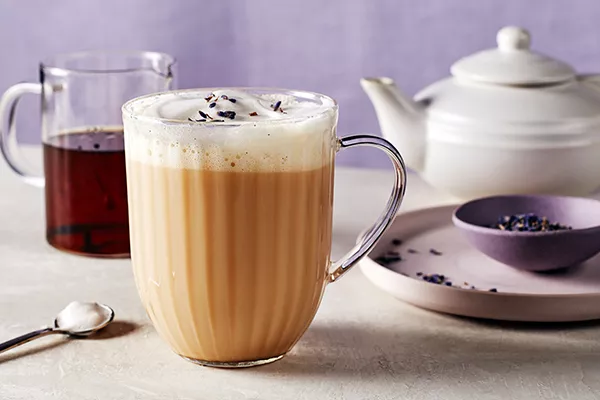  A glass mug of London Fog Latte with milk foam, shown with a pitcher of vanilla syrup, a tea pot, a stirring spoon, and a purple bowl of loose Earl Grey tea on a plate