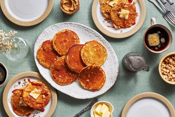 A table set with a platter of peanut butter pancakes, butter, a bowl of chopped peanuts, peanut butter, a mug of coffee, cutlery, a pitcher of syrup, and two plates with pancakes, syrup, and butter