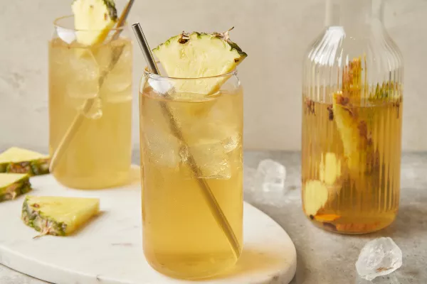 Two glasses of tepache with metal straws and garnished with pineapple wedges, shown with a pitcher of tepache