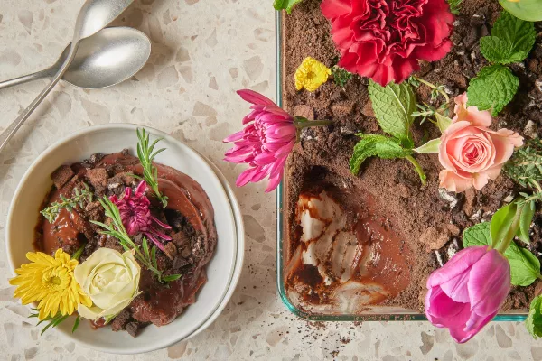Chocolate dirt pudding with cookie crumble topping garnished with edible flowers in a glass baking dish shown with a serving of pudding in a bowl with spoons