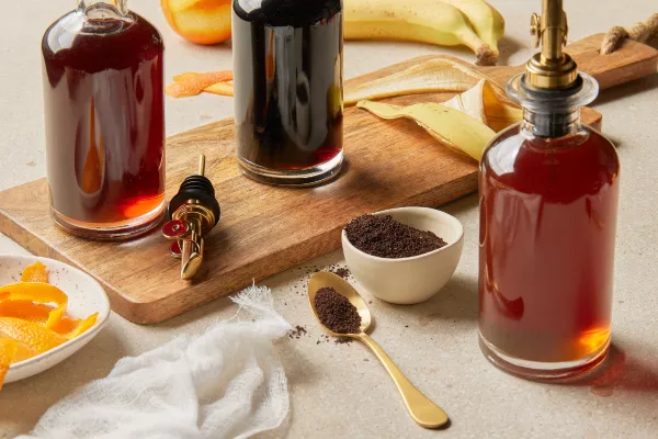 A bottle of coffee simple syrup, a bottle of orange simple syrup, and a bottle of banana simple syrup, shown on a table with a serving board, with coffee grounds, orange peels, and banana peels