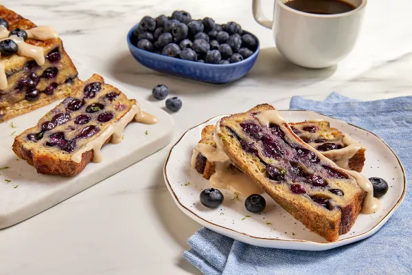 Two slices of loaf pan French toast with blueberries drizzled with maple glaze and shown with a cup of coffee, a dish of blueberries, and the partially sliced loaf 