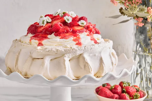 Pavlova topped with raspberries and strawberries and flowers on a white cake stand, shown with a vase of flowers and a bowl of berries