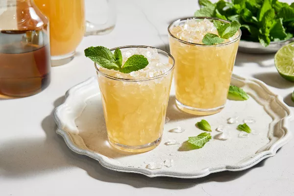 Two glasses of spirit-free mint julep with crushed ice and garnished with mint served on a white platter, shown with a bowl of mint leaves, half a lime, a pitcher of prepared julep and a pitcher of mint simple syrup