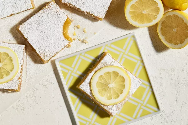 Lemon bars topped with icing sugar, two topped with a lemon slice, with one on a square plate with a yellow and white stripe pattern, shown with sliced lemons.