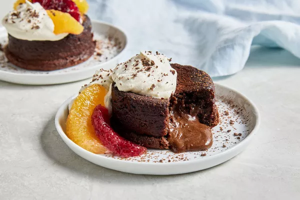 Two personal-size molten chocolate lava cakes on white plates, topped with whipped cream and cocoa powder, garnished with orange and blood orange segments, one shown with a piece missing and chocolate sauce spilling out from the centre.