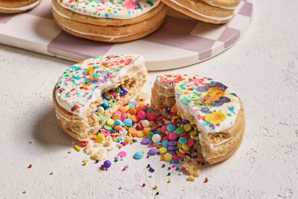 An iced pinata sugar cookie broken in two with candy confetti and sprinkles spilling out, shown with three more cookies on a white and mauve checkered tray in the background.