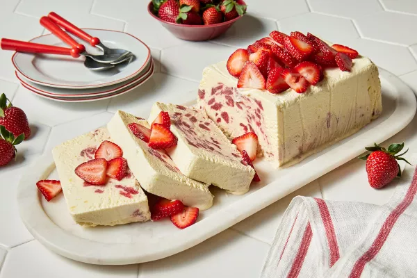 A rectangular loaf of strawberry-elderflower semifredo on a marble platter, garnished with sliced strawberries, with three slices cut, shown with three white plates with red trim, three red spoons, and a red bowl of strawberries.