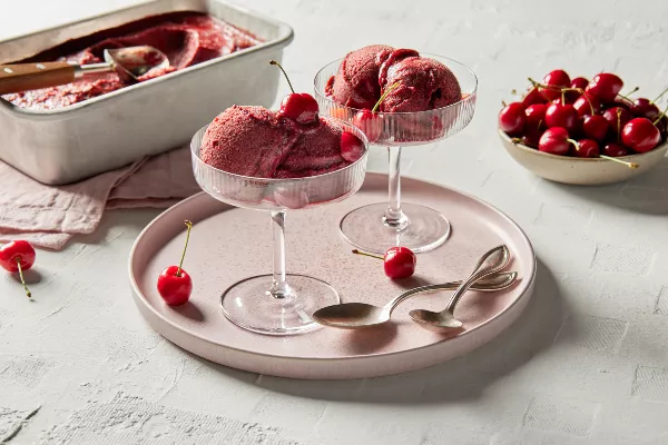 Two stemmed glass dishes of Cherry sorbet garnished with cherries, on a pale pink platter with gold spoons, shown with a bowl of cherries and a pan of cherry sorbet with a scoop for serving.